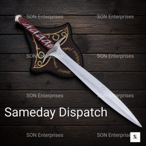 May include: A silver-toned sword with a pointed blade and a curved crossguard, featuring a handle with a red and black wood-grain pattern. The sword rests on a dark, shield-shaped plaque decorated with gold-coloured filigree and etched text.