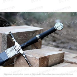 May include: A medieval sword with a black handle and silver detailing. The blade is in a dark scabbard. The hilt has an ornate design, and the pommel features a circular emblem. The sword is displayed on wooden blocks.