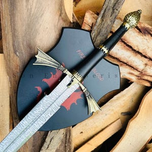 May include: A decorative sword with a patterned blade and gold-coloured accents. The black handle is wrapped in leather. A black shield with red dragon imagery and the words "Fire and Blood" is behind the sword.