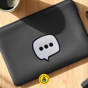 Op de afbeelding: Een zwarte laptoptas met een witte tekstballonafbeelding met drie zwarte stippen. De hoes heeft een ritssluiting en ligt op een houten oppervlak. Een pen, een kopje koffie en een smartphone zijn ook zichtbaar.