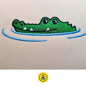 May include: An embroidered patch featuring a green crocodile head emerging from water. The crocodile has black outlines, white teeth, and black eyes. Below the crocodile, blue embroidered lines represent ripples in the water on a light beige fabric.