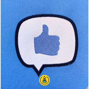 May include: Embroidered patch featuring a blue thumbs-up inside a white speech bubble with a black outline. The patch is on a light blue fabric background. A small yellow and blue circular emblem is in the lower corner.
