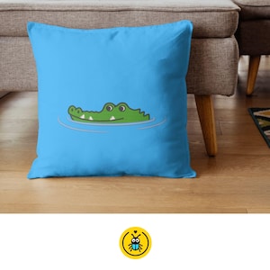 May include: A blue pillow with a green crocodile cartoon illustration on it. The crocodile is swimming in water and has a toothy grin.