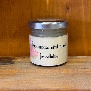 Op de afbeelding: Een klein glazen potje met een zilveren deksel dat een witte zalf bevat. Het etiket vermeldt "Bijenwaszalf voor cellulitis" met roze rozen op het etikett.