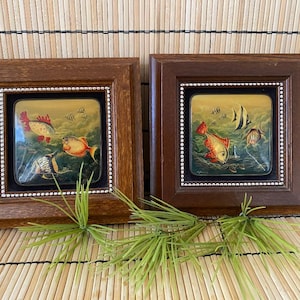 May include: Two framed square artworks featuring colorful fish swimming in an underwater scene. The frames are dark brown with a beaded inner border. The fish are orange, red, and yellow, set against a green and blue background.