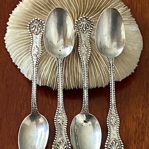 May include: Four antique silver spoons with ornate handles and oval bowls are displayed against a cream-colored, textured background. The spoons have detailed floral and scrollwork designs. The spoons are arranged vertically, showcasing their elegant craftsmanship.