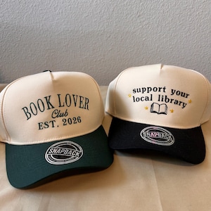 Peut inclure: Deux casquettes de camionneur à fermeture à pression. L'une est crème et vert forêt avec "BOOK LOVER CLUB EST. 2026" brodé sur le devant. L'autre est crème et noire avec "support your local library" brodé.