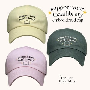 May include: Three baseball caps in pale yellow, light pink, and dark green. Each cap features embroidered text that reads support your local library, accompanied by a small open book icon and four small star accents centered on the front panel.