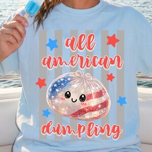 May include: Light blue t-shirt with the text "all american dumpling" and a graphic of a dumpling decorated with the American flag. The shirt is paired with red and white checkered shorts. A red, white, and blue popsicle is held.