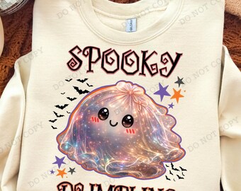 May include: A cream-colored crewneck sweatshirt featuring a graphic of a galaxy-patterned dumpling character with a smiling face. The text "SPOOKY DUMPLING" is printed in a dark font above and below the character, surrounded by small bats and stars.