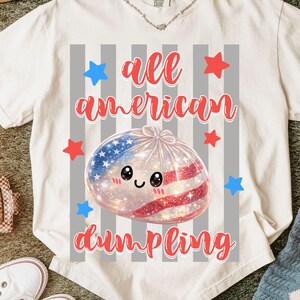 May include: White t-shirt with the text "all american dumpling" in red script. The graphic features a dumpling with an American flag design, surrounded by red and blue stars. The shirt is on a green surface.