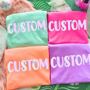 May include: Four folded t-shirts in shades of green, pink, orange, and purple. Each shirt has the word "CUSTOM" printed in a playful font with a polka dot and striped design. The shirts are arranged on a colorful background.
