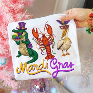 May include: White t-shirt featuring embroidered Mardi Gras designs. Includes a green alligator with a purple hat, an orange crawfish, and a pelican wearing a mask. The words "Mardi Gras" are written in yellow and purple.