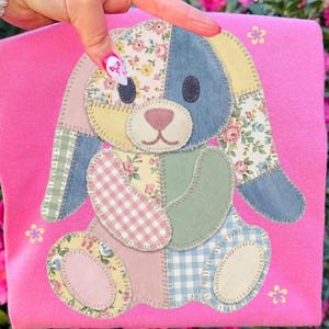 Może przedstawiać: Różowa koszulka z patchworkowym wzorem królika. Królik wykonany jest z różnych wzorzystych tkanin, w tym kwiatowych, w kratkę i jednolitych kolorów. Wzór jest naszyty na koszulce, z małymi kwiatowymi akcentami.