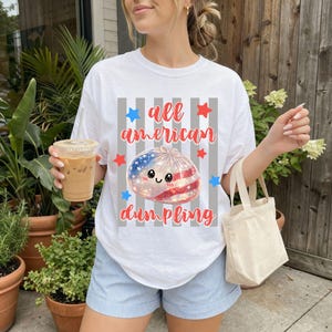 May include: White t-shirt with the text "all american dumpling" in red script. The graphic features a dumpling with an American flag design, surrounded by red and blue stars. The shirt is worn with blue shorts.