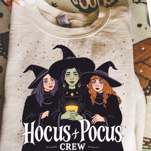 Peut inclure: Un sweat-shirt beige avec un motif de trois sorcières portant des chapeaux et des robes noirs, avec le texte "Hocus Pocus Crew". Les sorcières ont des couleurs de cheveux différentes et tiennent une bougie allumée.