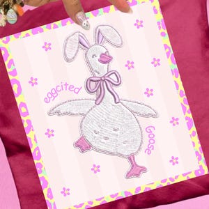 May include: A square print with a pink and white striped background, featuring an embroidered goose wearing bunny ears and a bow. The text "eggcited Goose" is written on the print. The print has a leopard print border.