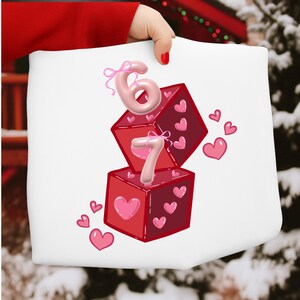 May include: White tote bag featuring a design with red dice, pink hearts, and the numbers 6 and 7. The numbers are in a pink, balloon-like font, and the dice have heart details. The design is perfect for Valentine's Day.