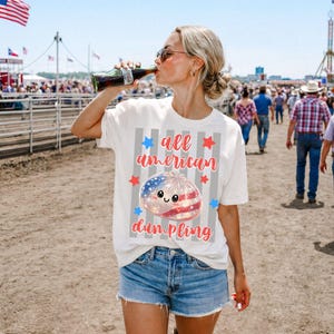May include: White t-shirt with the text "all american dumpling" and a graphic of a dumpling with an American flag design. The shirt is paired with denim shorts. A person is drinking from a Coca-Cola bottle.