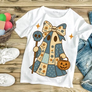 Op de afbeelding: Wit T-shirt met een grillig spookontwerp. Het spook draagt een patchwork blauw en beige outfit met bloemaccenten, een schedelstrik en een pompoenemmer. Het houdt een staf met een schedel vast. Halloween-kleding.
