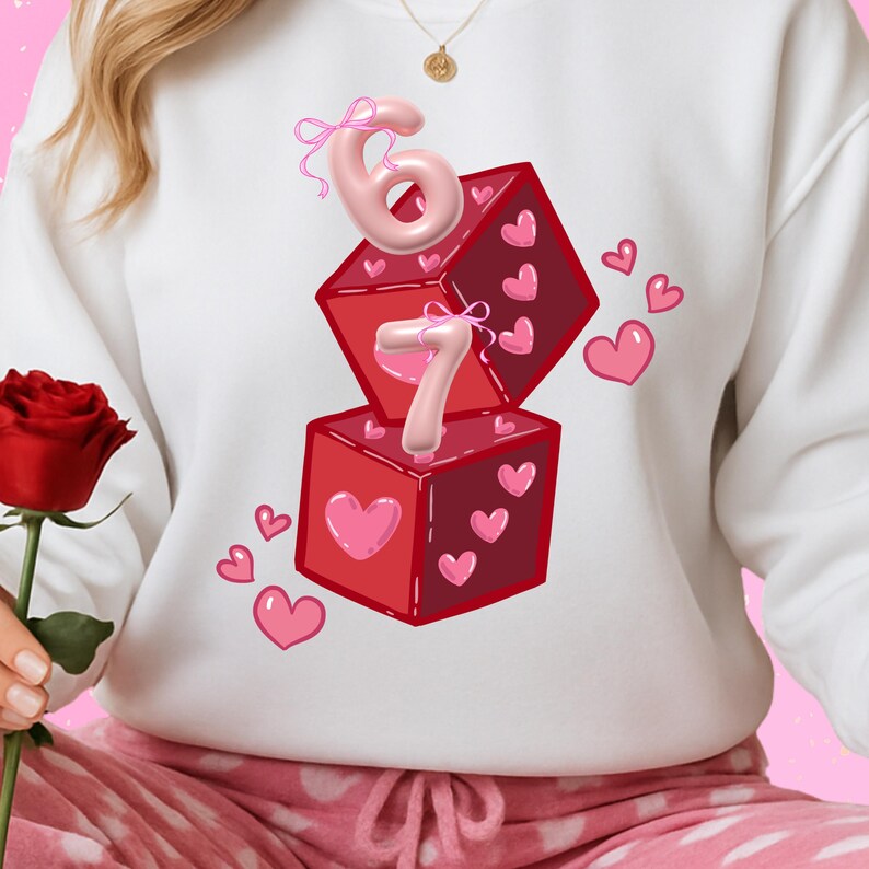 May include: White sweatshirt featuring a design of two red dice with pink hearts and the numbers 6 and 7. The numbers are in a pink, 3D style. Pink hearts are scattered around the dice.