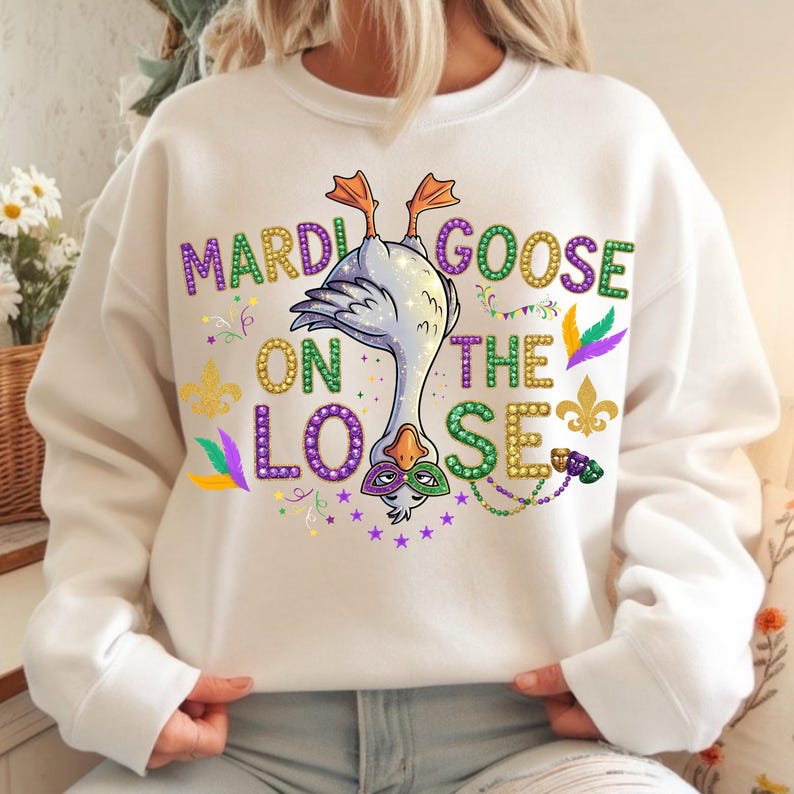 May include: White sweatshirt with a whimsical design featuring a goose wearing a mask, with the text "Mardi Goose on the Loose" in purple, green, and gold. The design includes stars, feathers, and fleur-de-lis elements, perfect for Mardi Gras celebrations.