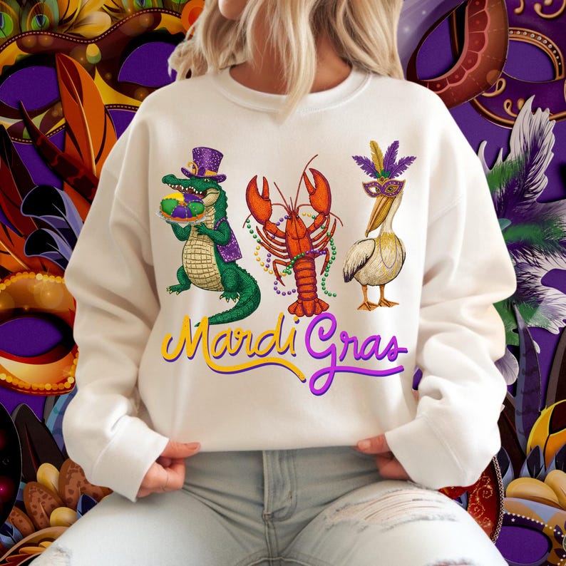 May include: Cream-colored sweatshirt featuring a Mardi Gras design. The graphic includes an alligator, a lobster, and a pelican, all adorned with festive accessories. The words "Mardi Gras" are written in a colorful script across the bottom.