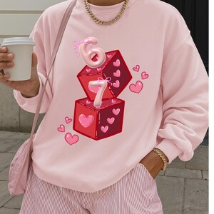 May include: A light pink sweatshirt with a graphic of two red dice with heart details and the numbers 6 and 7. Pink hearts are scattered around the dice. The sweatshirt is part of a casual clothing collection.