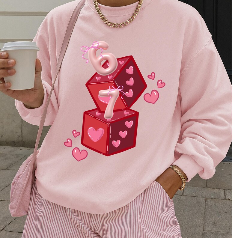 May include: A light pink sweatshirt with a graphic of two red dice with heart details and the numbers 6 and 7. Pink hearts are scattered around the dice. The sweatshirt is part of a casual clothing collection.