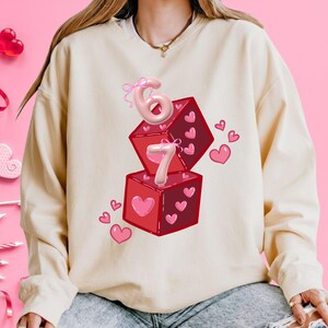 May include: Cream-colored sweatshirt featuring a graphic of two red dice with pink hearts and the numbers 6 and 7. The numbers are pink and appear to be made of balloons. Small pink hearts are scattered around the dice.