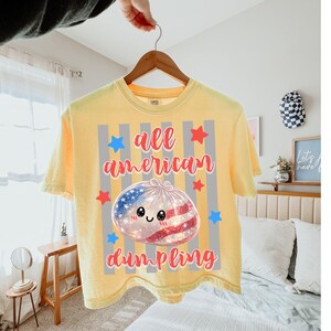 May include: Yellow t-shirt with the text "all american dumpling" in red script. The graphic features a dumpling with an American flag design and a cute face, surrounded by stars and stripes.