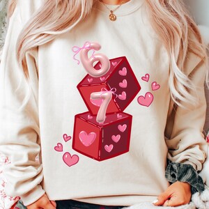 May include: Cream-colored sweatshirt featuring a graphic of two red dice with pink hearts and the numbers 6 and 7. The numbers are in a 3D style with pink bows. Pink hearts are scattered around the dice.