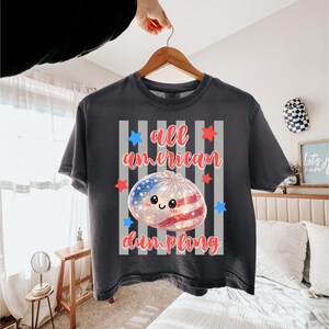 May include: Dark gray t-shirt with the text "all american dumpling" in red script. The design features a dumpling with a smiling face, and a red, white, and blue American flag design. The shirt is hanging on a wooden hanger.