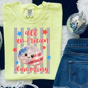 May include: A light yellow Comfort Colors t-shirt with the words "all american dumpling" in red script. The graphic features a dumpling with an American flag design, surrounded by red and blue stars.