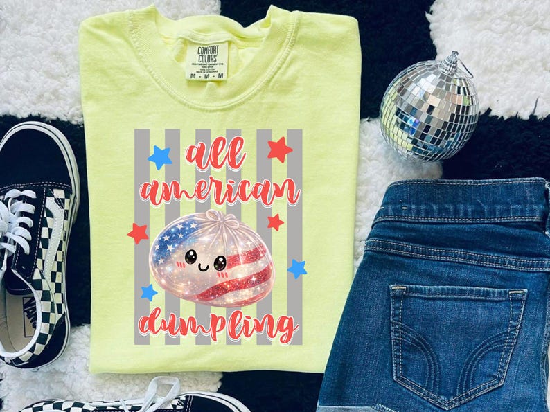 May include: A light yellow Comfort Colors t-shirt with the words "all american dumpling" in red script. The graphic features a dumpling with an American flag design, surrounded by red and blue stars.