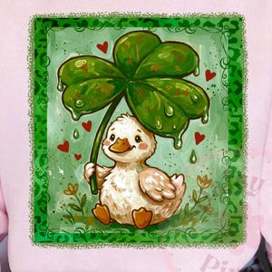 May include: A light pink sweatshirt with a St. Patrick's Day graphic. The graphic shows a duckling holding a large green four-leaf clover, with red hearts and green accents. The duckling is white with brown feet.