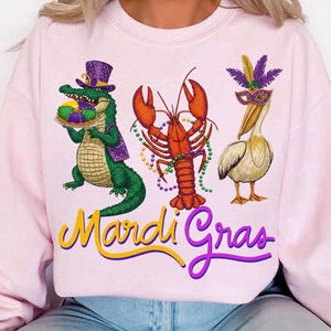 May include: Pink sweatshirt featuring a colorful Mardi Gras design. The graphic includes an alligator in a purple hat, a red lobster, and a pelican wearing a mask. The words "Mardi Gras" are written in yellow and purple.