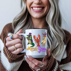 May include: White ceramic mug featuring Mardi Gras themed illustrations of an alligator, lobster, and pelican. The mug has the words "Mardi Gras" in yellow and purple script. The mug is held by a person.