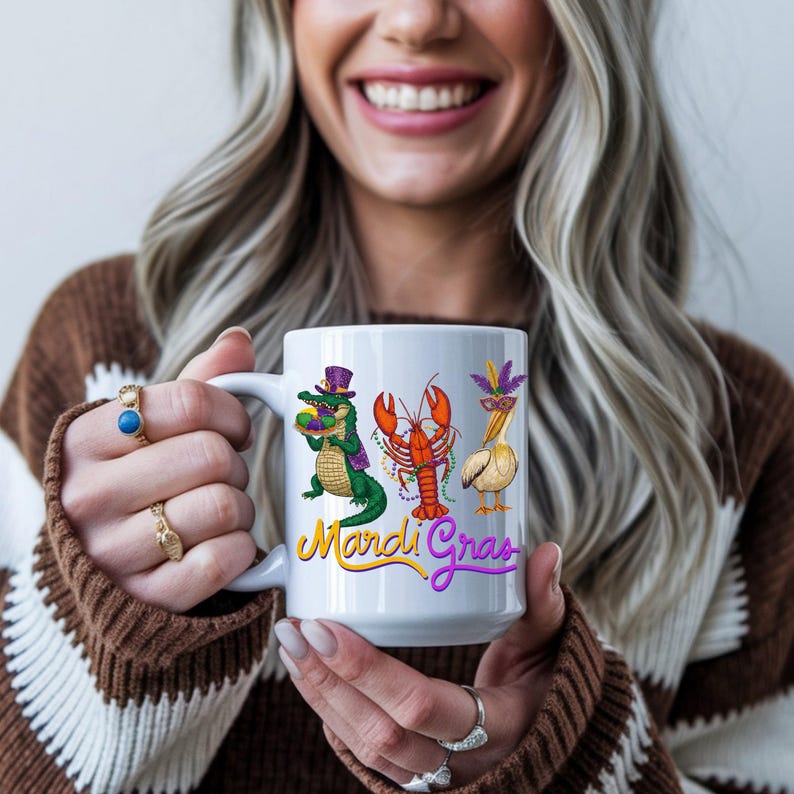 May include: White ceramic mug featuring Mardi Gras themed illustrations of an alligator, lobster, and pelican. The mug has the words "Mardi Gras" in yellow and purple script. The mug is held by a person.