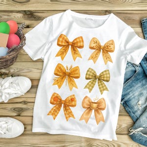 Puede incluir: Camiseta blanca con seis diseños de lazo de acuarela en tonos naranja, amarillo y verde. Los lazos tienen patrones como cuadros y florales, con detalles de calabaza y taza de café. La camiseta se muestra sobre una superficie de madera.