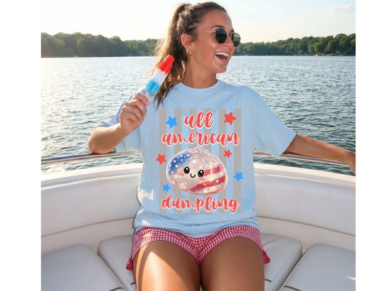 May include: Light blue t-shirt with the text "all american dumpling" and a graphic of a dumpling decorated with the American flag. The shirt is paired with red and white checkered shorts. A red, white, and blue popsicle is held.