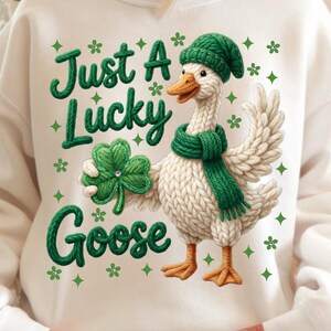 Może przedstawiać: Biała bluza z nadrukiem gęsi w zielonym kapeluszu i szaliku, trzymającej koniczynę. Tekst "Just A Lucky Goose" jest zielony, z zielonymi akcentami koniczyny i diamentów.