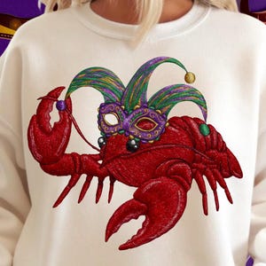 Puede incluir: Sudadera color crema con un diseño bordado. Un cangrejo rojo está adornado con una máscara de bufón morada y verde. El diseño es una representación festiva del Mardi Gras, ideal para celebraciones.