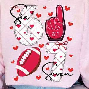 May include: Pink sweatshirt with a design featuring the numbers 6 and 7, a football, and a foam finger. The numbers are white with a red heart pattern. The word "Six" and "Seven" are written in black script, with red hearts scattered around.