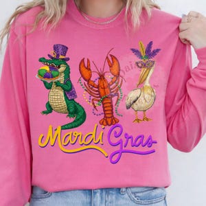 May include: Pink long-sleeve shirt with a Mardi Gras theme. Features an alligator, lobster, and pelican with festive accessories. The words "Mardi Gras" are written in yellow and purple script.