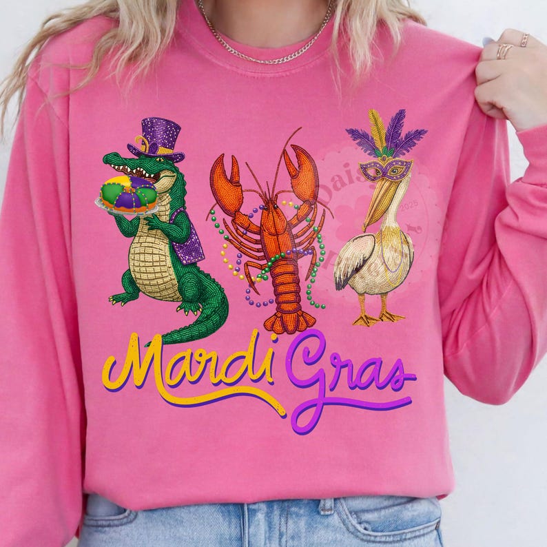 May include: Pink long-sleeve shirt with a Mardi Gras theme. Features an alligator, lobster, and pelican with festive accessories. The words "Mardi Gras" are written in yellow and purple script.