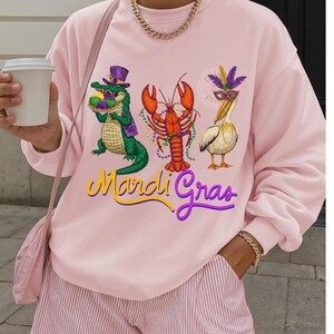May include: A light pink sweatshirt featuring Mardi Gras themed illustrations. The design includes a green alligator wearing a purple hat, an orange lobster, and a white pelican wearing a mask. The words "Mardi Gras" are written in yellow and purple.