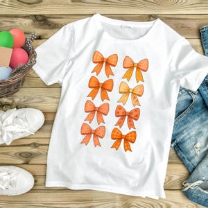 Puede incluir: Camiseta blanca con un diseño de ocho lazos naranjas en varios diseños. Los lazos son en tonos naranja y melocotón. La camiseta está sobre una superficie de madera, con una cesta de huevos coloridos y zapatillas blancas.