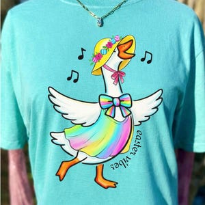 May include: A turquoise t-shirt with a cartoon goose design. The goose wears a yellow hat with flowers, a rainbow dress, and a pink bow. Musical notes surround the goose, and the words "easter vibes" are written on the shirt.