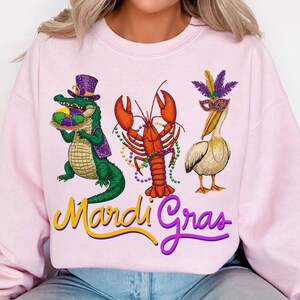 May include: A light pink sweatshirt featuring a colorful Mardi Gras design. The design includes a green alligator wearing a purple hat, an orange lobster, and a white pelican wearing a mask. The words "Mardi Gras" are written in yellow and purple.
